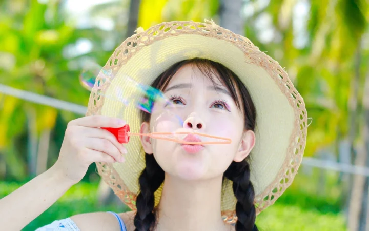 麦わら帽子をかぶった若い女性が、緑豊かな屋外でシャボン玉を吹いている。