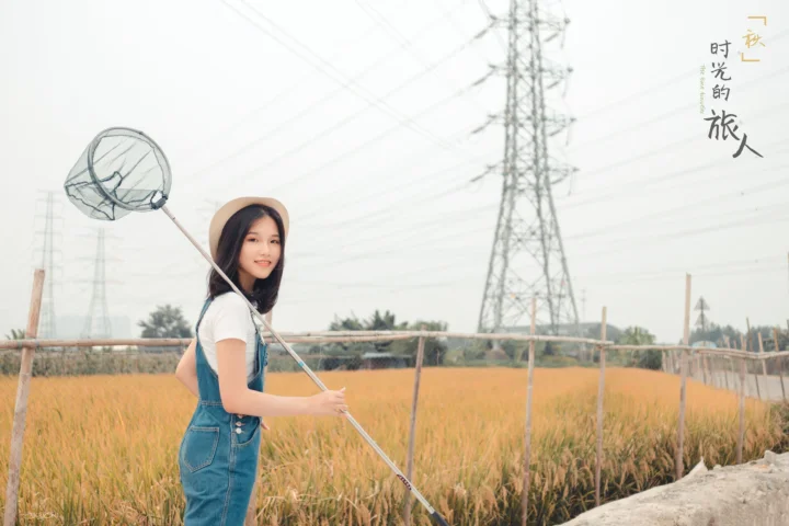 一名穿白色上衣和蓝色背带裤的年轻女子站在麦田边，手持捕虫网，背景是电塔与晴朗天空。