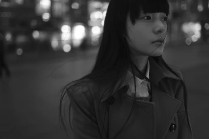一名穿着深色外套的女性站在背景虚化的街道上，画面采用黑白色调。