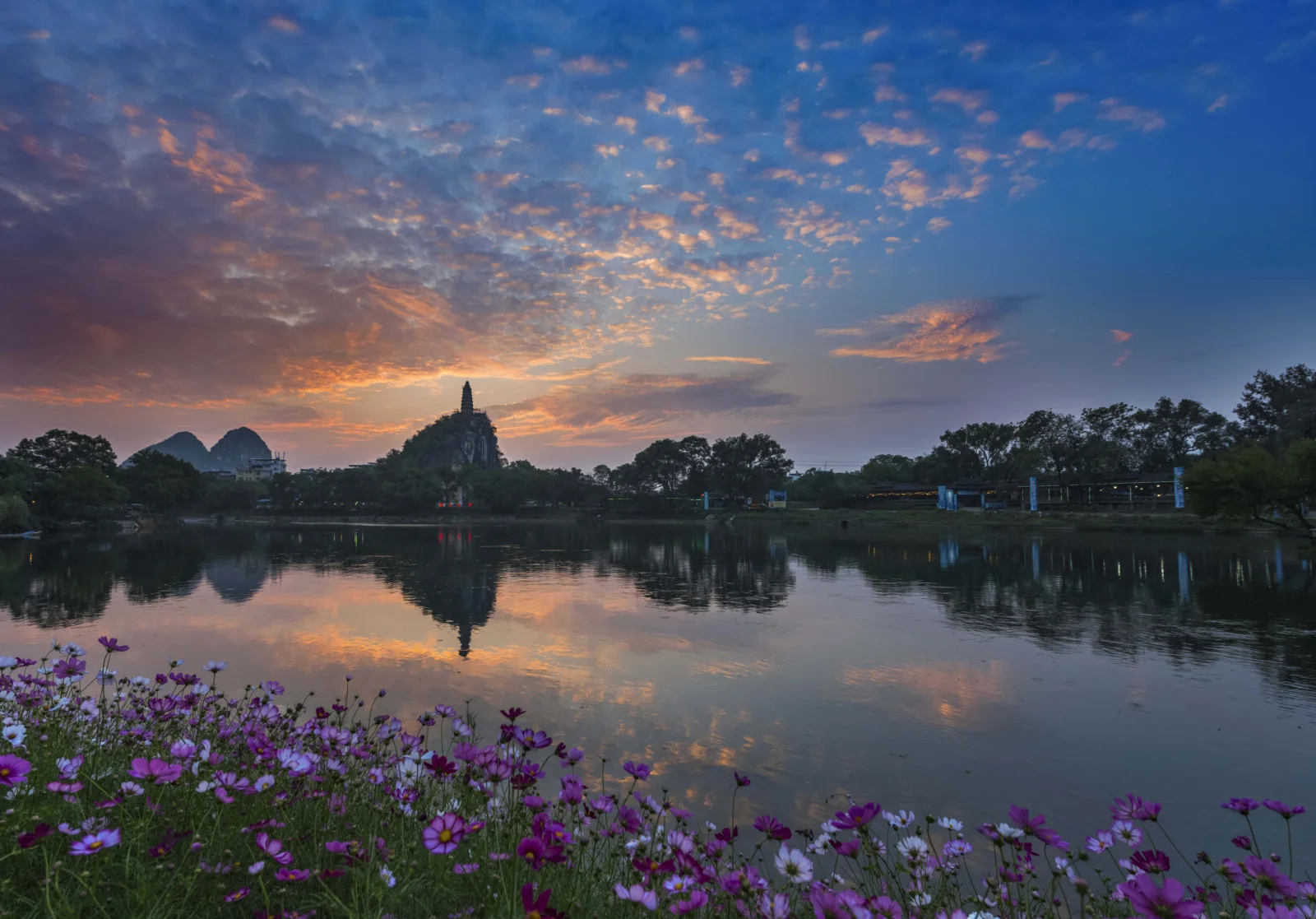 夕阳映照湖面，远处山峦与尖塔倒映在水中，岸边盛开着紫色花朵。