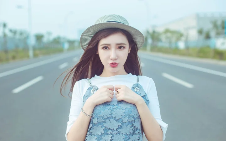 一名戴着宽檐帽的女性站在宽阔的公路上，双手握着蓝色蕾丝连衣裙的肩带。