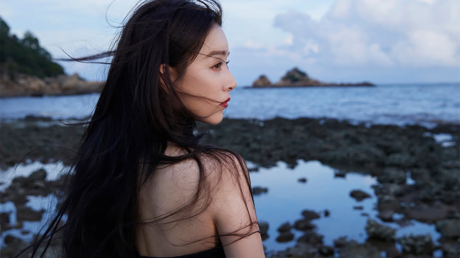 一名成年女性站在海邊岩石岸旁，側臉望向平靜的海面。