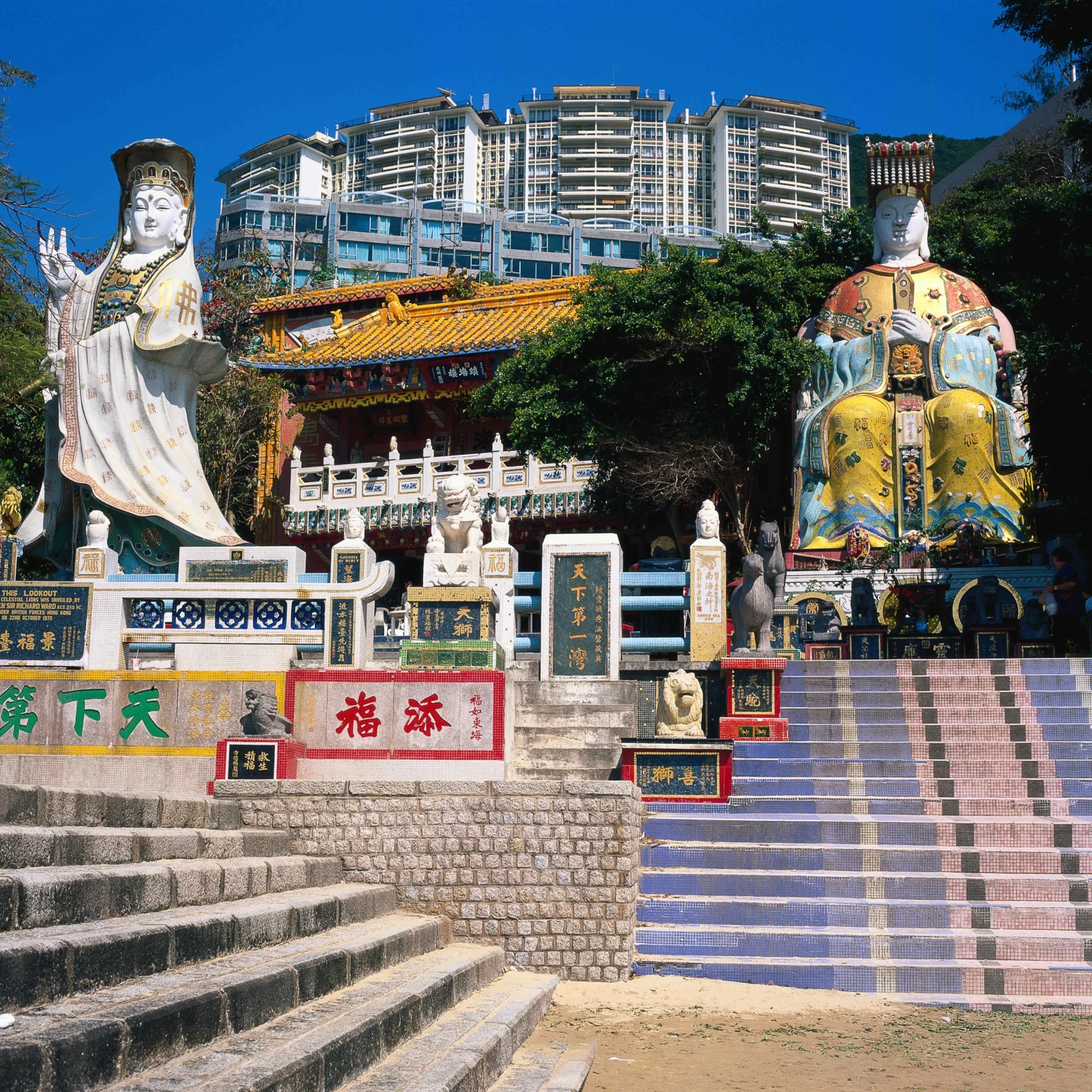 阳光下的寺庙庭院中立着两尊大型彩色雕像，后方是现代城市建筑。