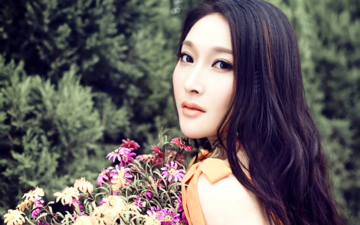 一名长发女性身穿橙色无袖上衣，置身于彩色花朵和绿色树林前。
