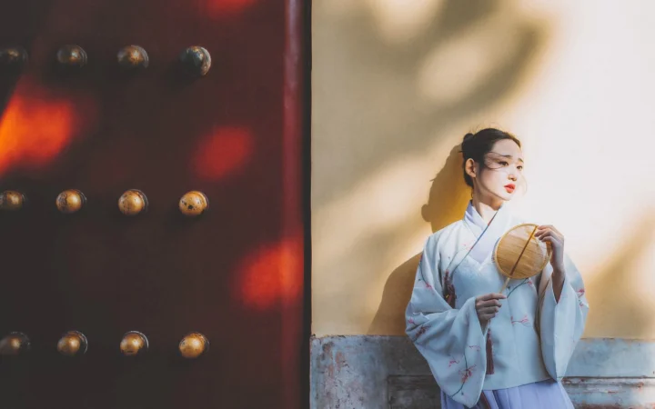 一名身穿浅色传统服饰的女子站在木门与浅色墙面旁，手持圆扇，阳光投下柔和的影子。
