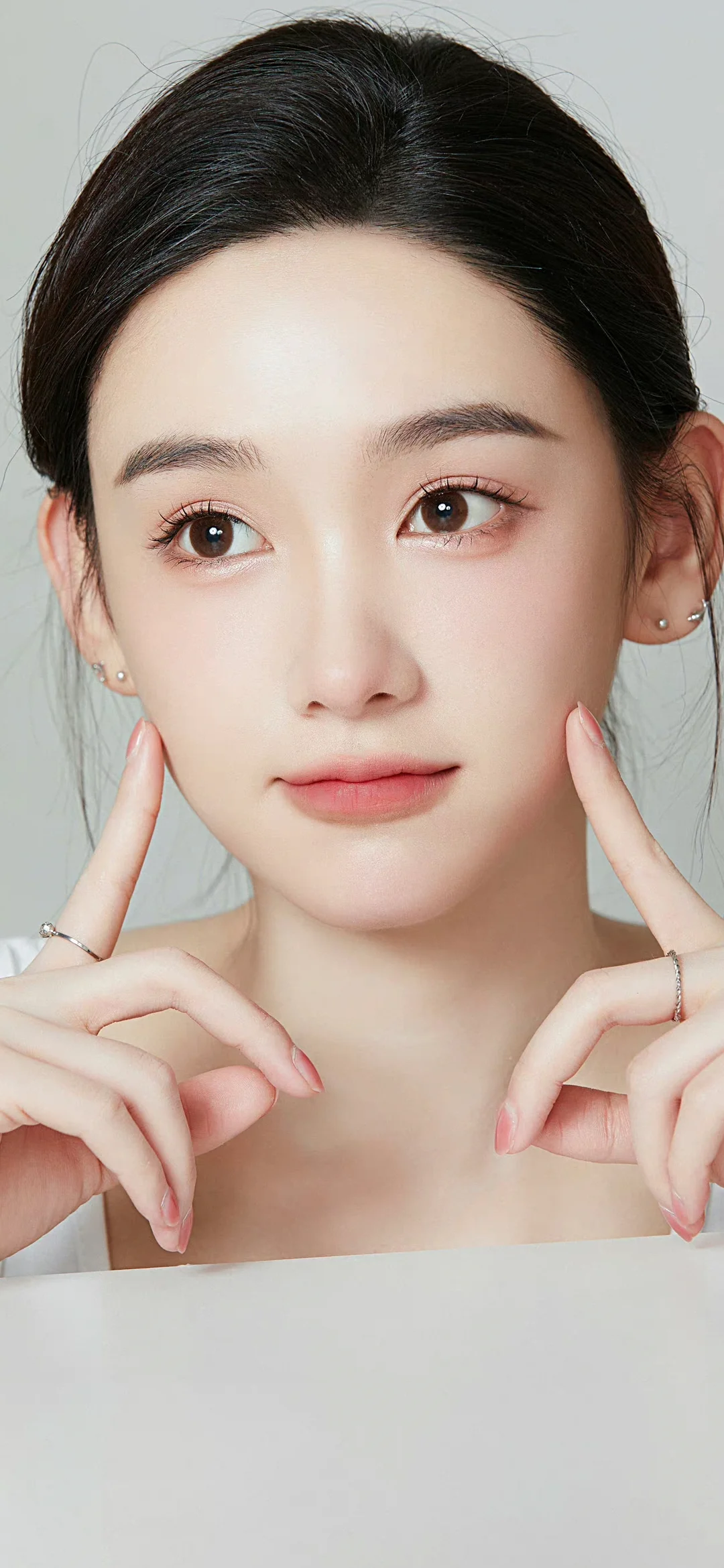 연한 회색 배경 앞에서 성인 여성이 양손을 볼에 대고 카메라를 바라보고 있습니다.