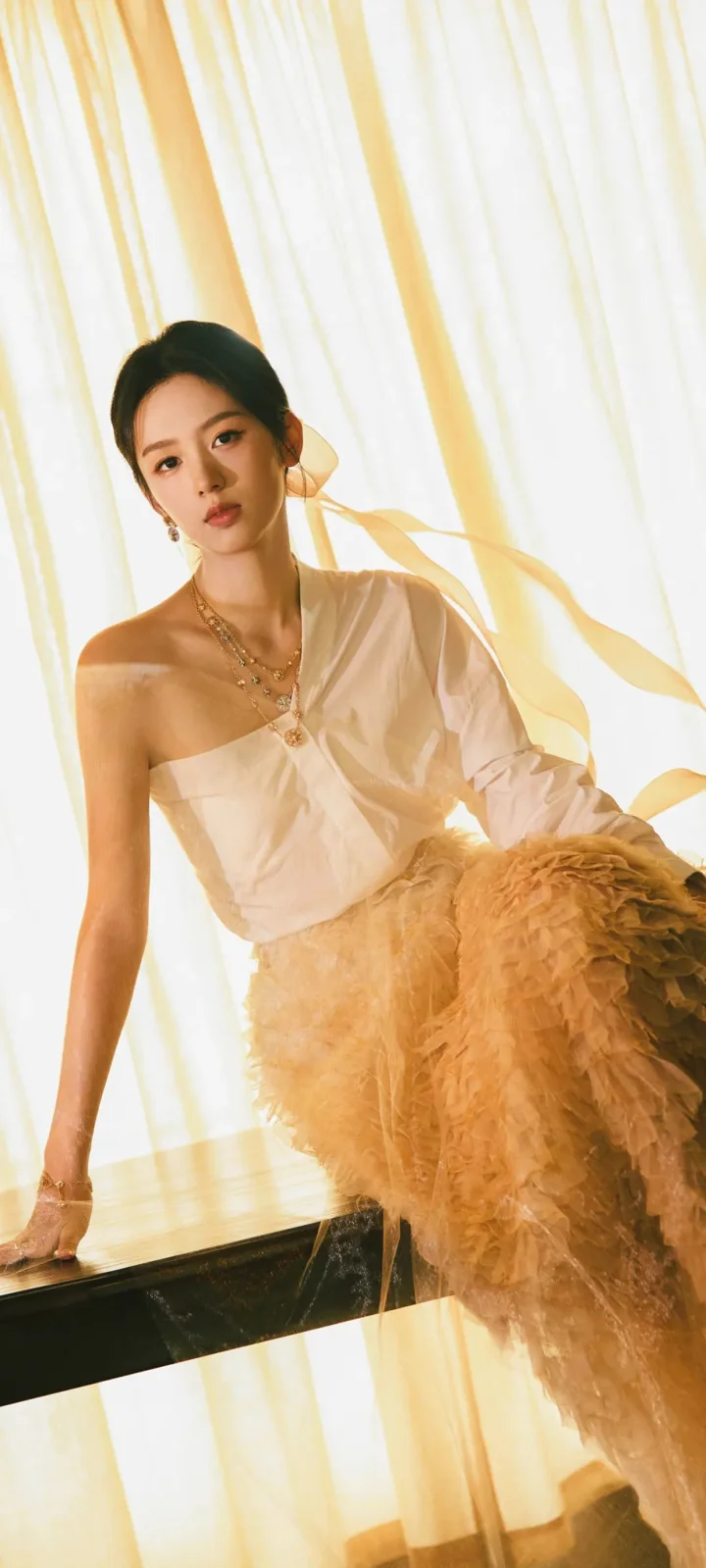 一位成年女性在暖色窗帘前身穿白色单肩服装，呈现时尚人像画面。