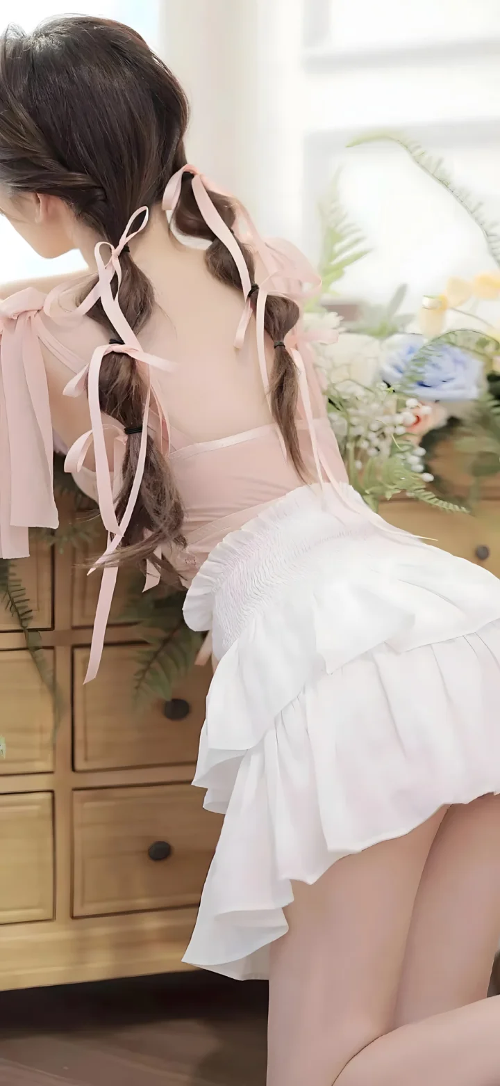 長い三つ編みの女性がピンクのトップスと白いフリルスカートを着て、木製のチェストと花束のそばに立っています。