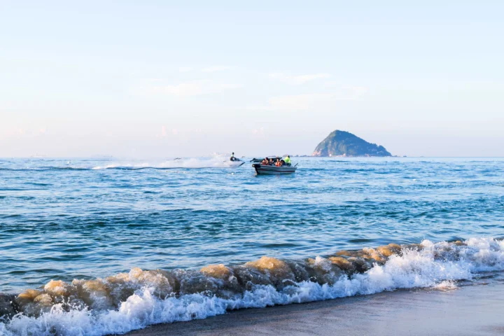 蓝色海面上漂浮着一只小船，远处可见海岛，前景是拍岸的浪花。