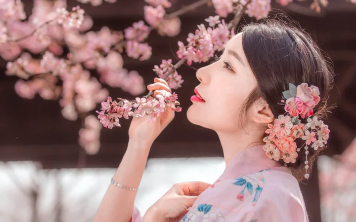 一位成年女性手持樱花枝，发间佩戴花朵饰品，置身粉色樱花之间。