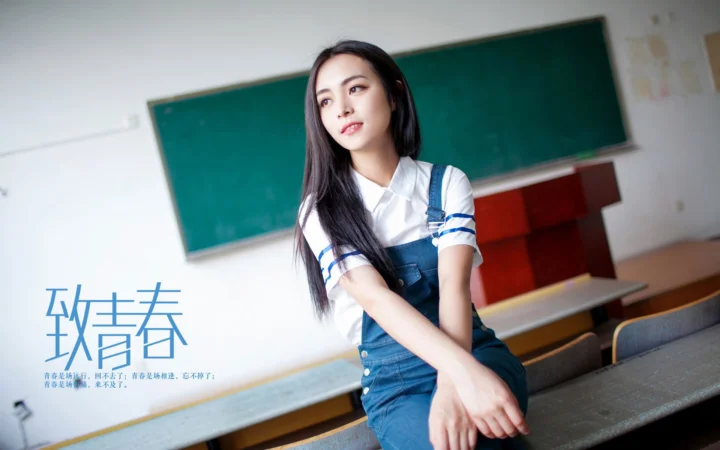 一名成年女性身穿白衬衫和蓝色背带裙坐在教室里，身后是黑板。