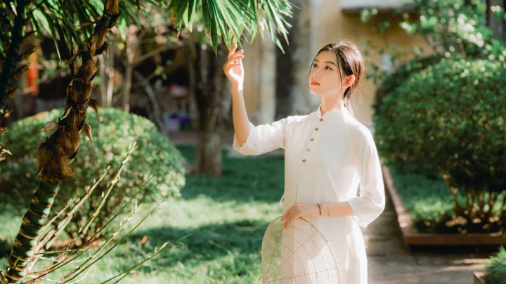 一名身穿白色长袖衣物的女子站在绿意盎然的庭院中，伸手触碰垂下的竹叶。