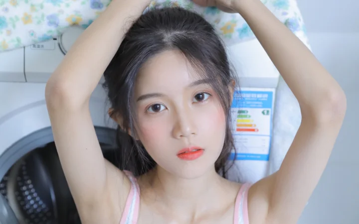 一名黑发女子抬起双臂，面向镜头凝视，背景可见室内设备与浅色床品。