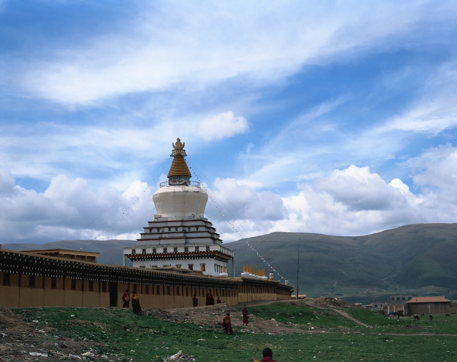一座白色佛塔矗立在群山与蓝天之间，周围环绕着寺院建筑和草地。