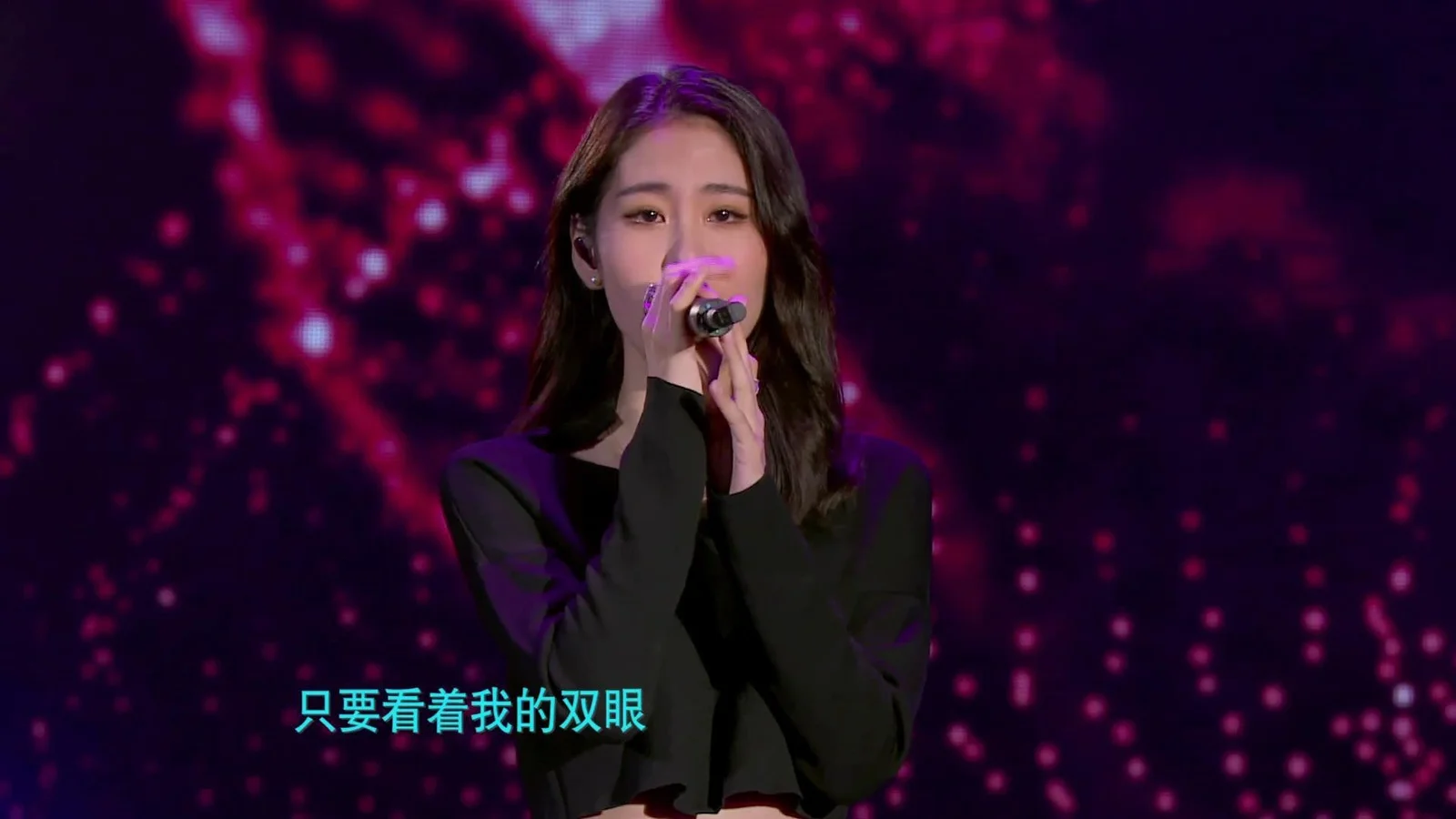 一位身穿黑色上衣的女歌手手持麦克风站在紫红色灯光背景前演唱。