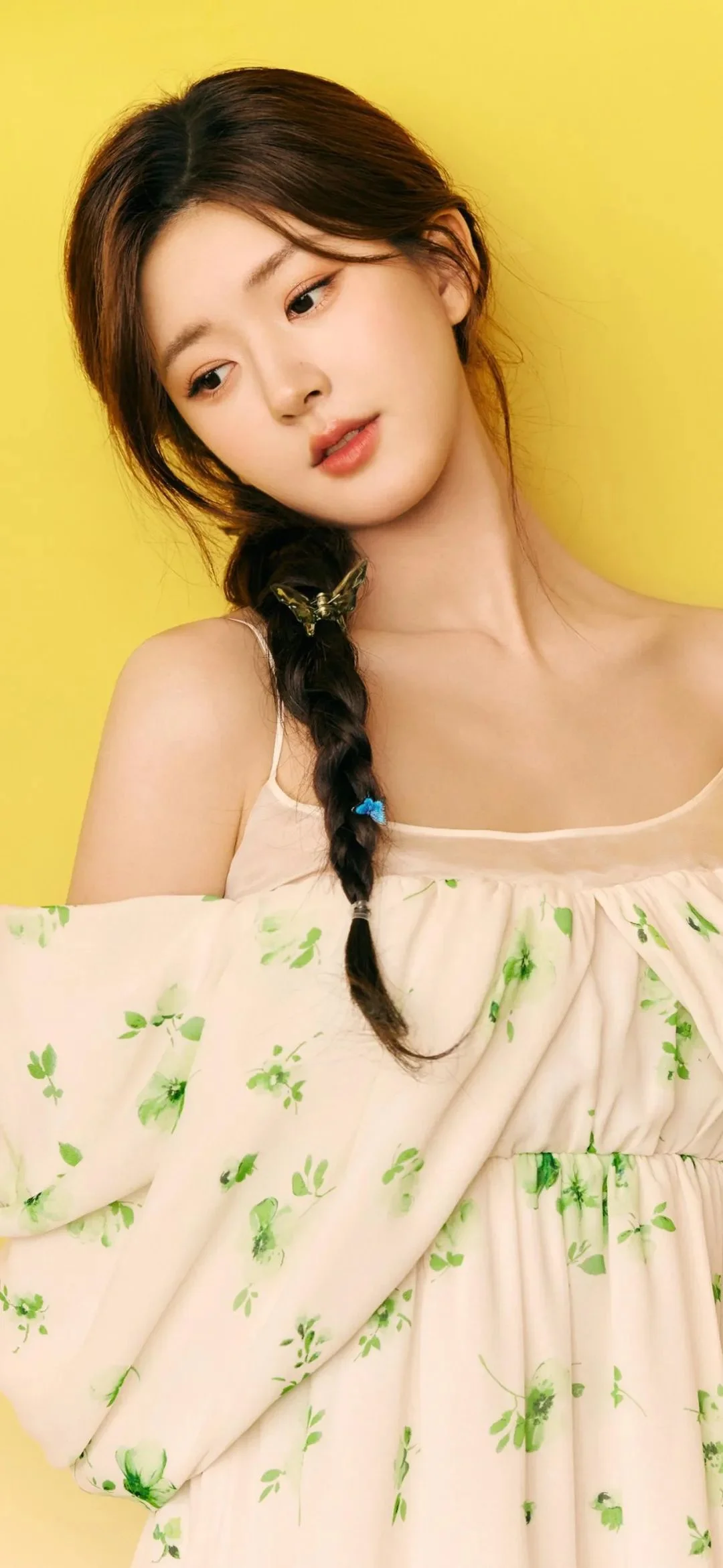 긴 땋은 머리의 여성이 연한 꽃무늬 원피스를 입고 밝은 노란색 배경 앞에 서 있다.