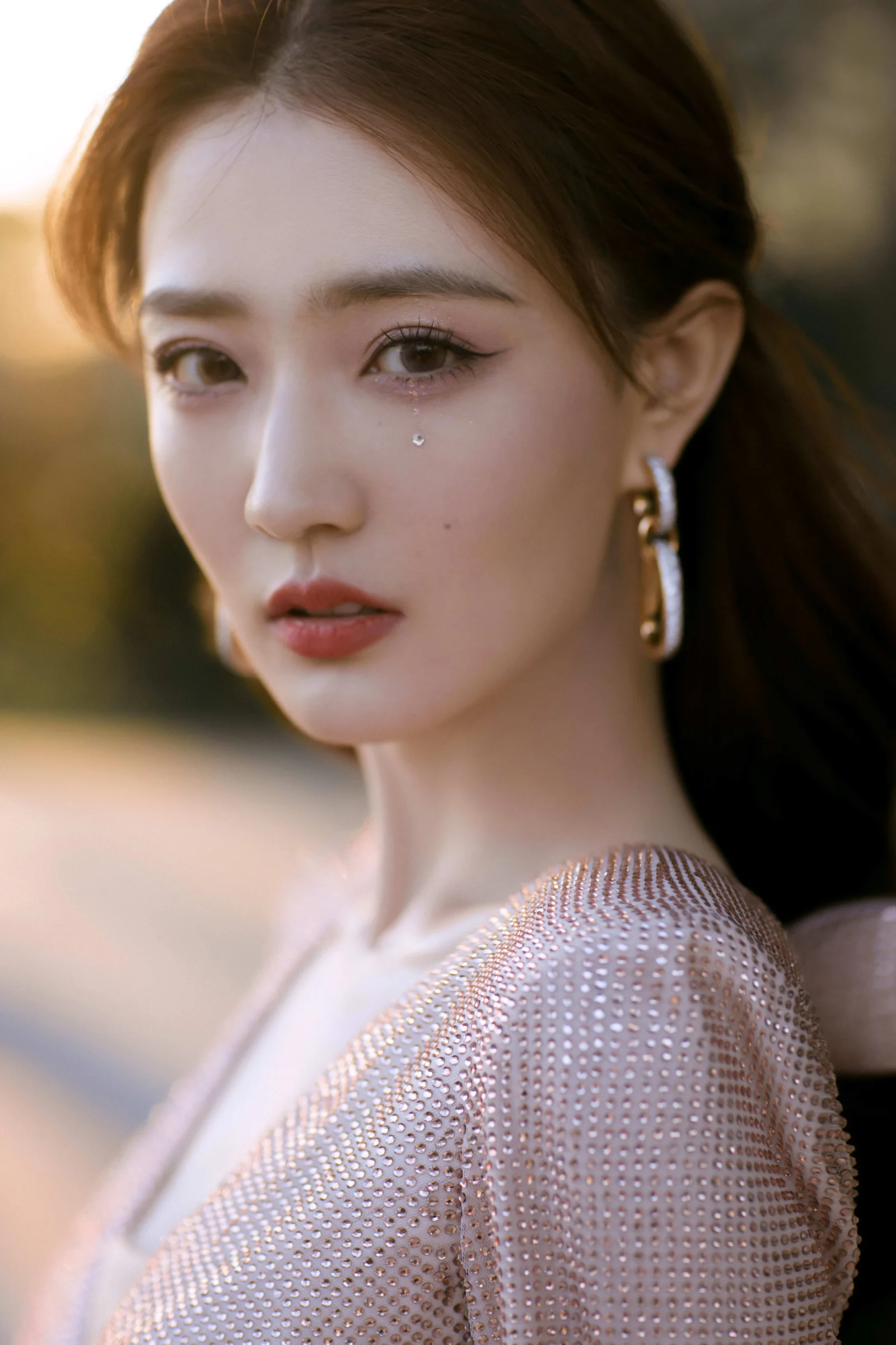 一位佩戴长耳环、身穿闪亮上衣的女性在暖色虚化背景前凝视镜头。