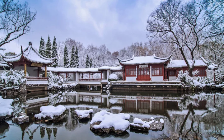 覆雪的中式园林建筑坐落在池塘边，静谧水面映出冬日景色。