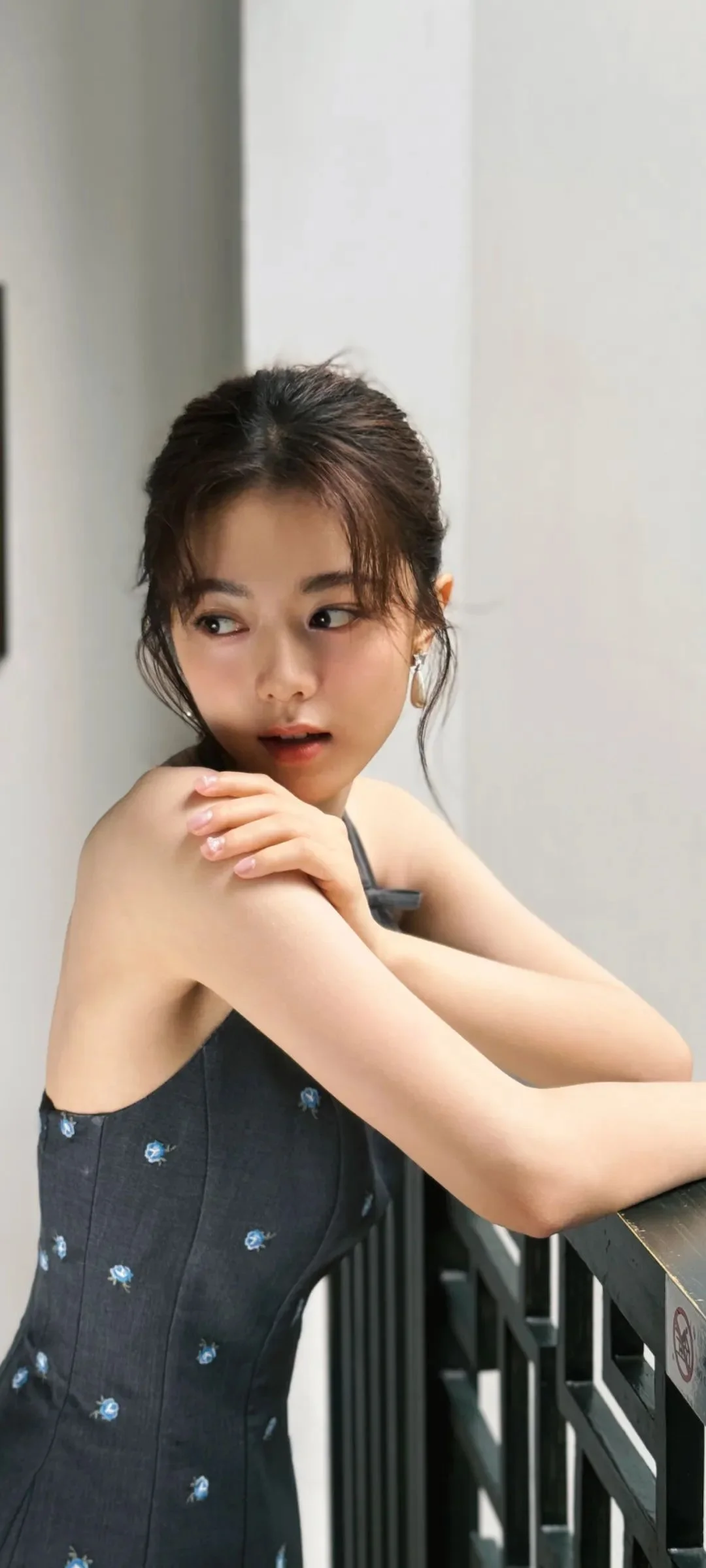 검은 머리의 여성이 검은 민소매 원피스를 입고 발코니 난간에 기대어 있다.