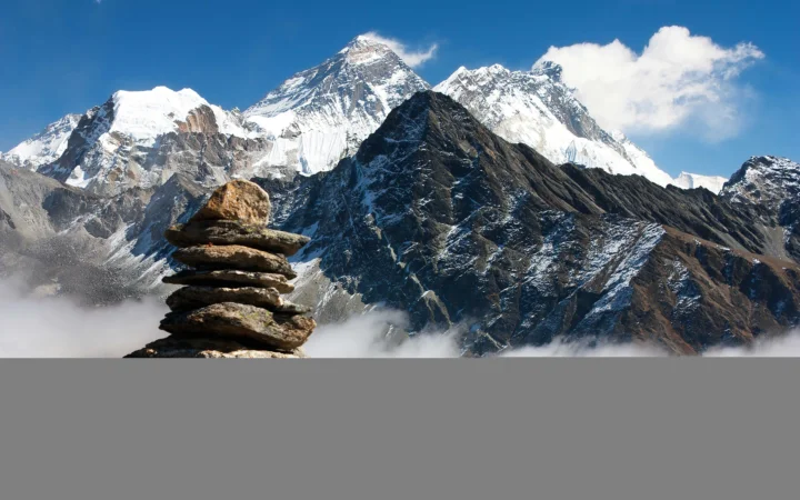 雄伟的雪山矗立在蓝天下，前景可见叠放的石块与山间云雾。