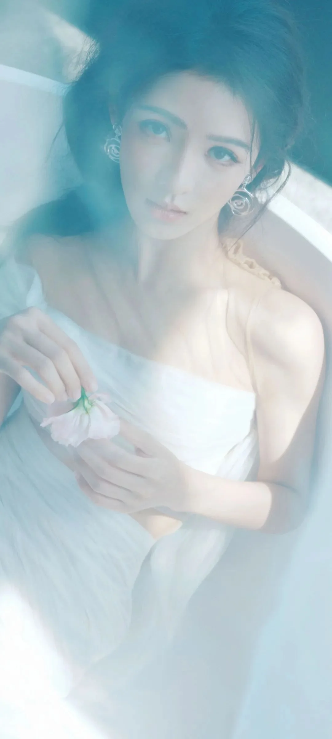 흰색 오프숄더 드레스를 입은 성인 여성이 연한 색의 꽃을 들고 부드러운 청백색 안개 속에서 카메라를 바라보고 있다.