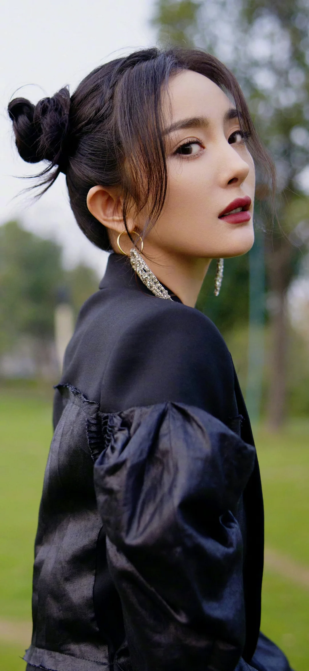 一名成年女性在户外回眸看向镜头，身穿黑色外套并佩戴长耳饰。