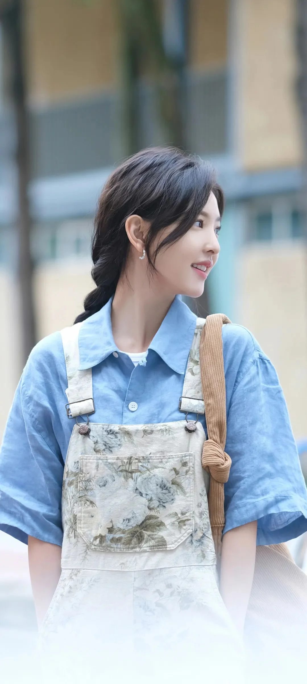 파란 셔츠와 꽃무늬 멜빵바지를 입은 성인 여성이 흐릿한 건물 배경 앞에 옆모습으로 서 있습니다.