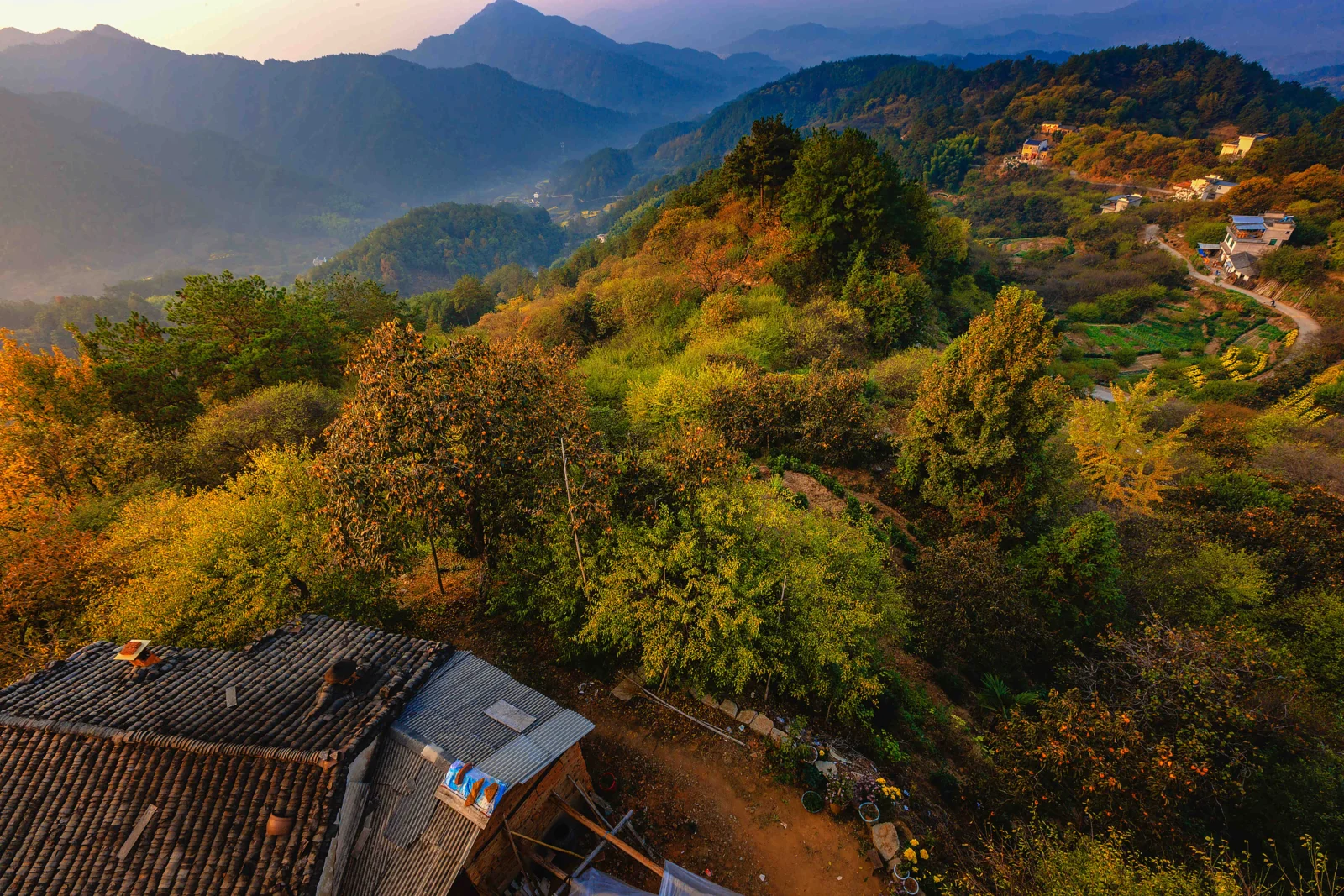 秋色山林环绕着山间村落，远处层叠群山延伸至天际，前景可见传统屋顶。