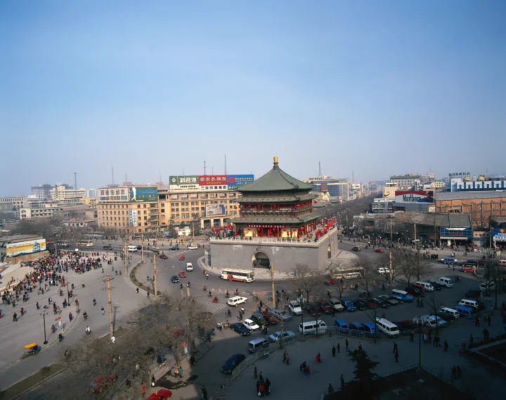 一座古建筑矗立在繁忙的城市广场中央，周围环绕着街道、车辆和行人。