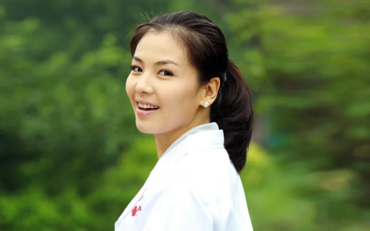 一名微笑的成年女性身穿白色上衣站在绿意盎然的户外环境中。