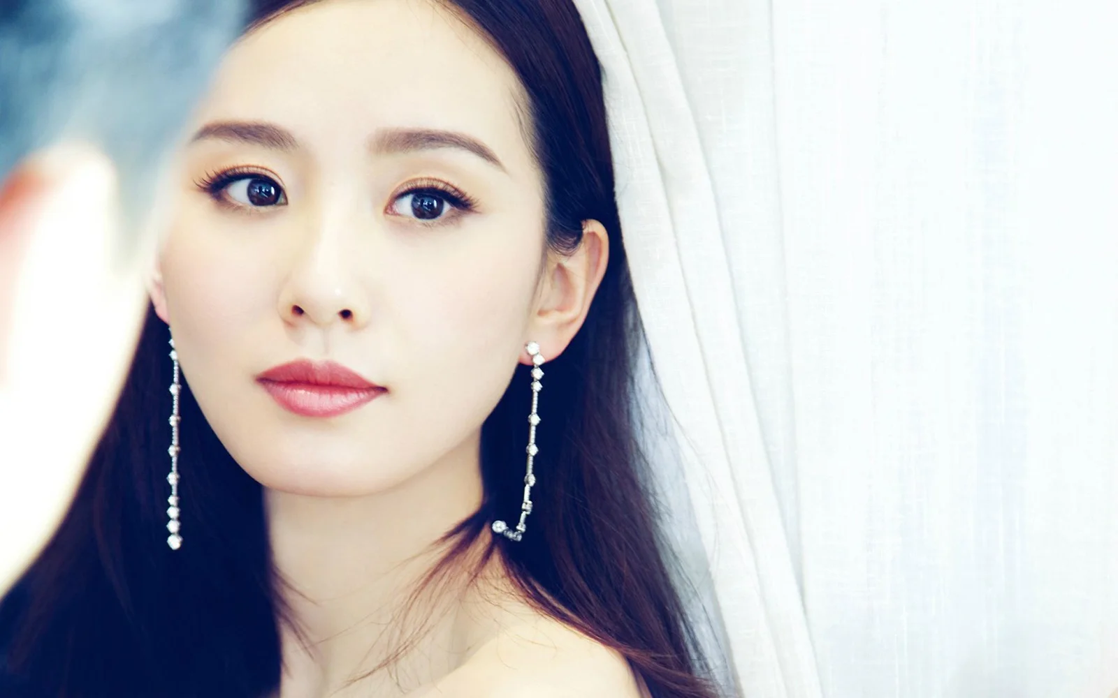 一位戴着垂坠耳环的成年女性在浅色窗帘前进行近距离肖像拍摄。