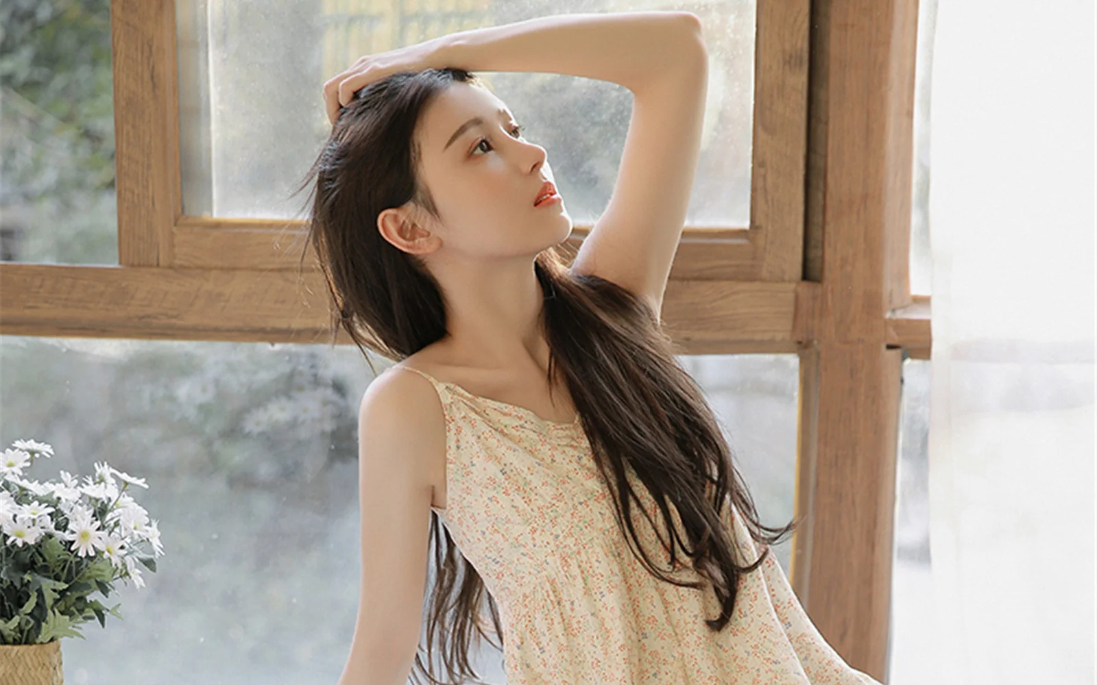 一位长发女子穿着碎花裙站在木框窗前，柔和的光线照亮了她的侧脸。