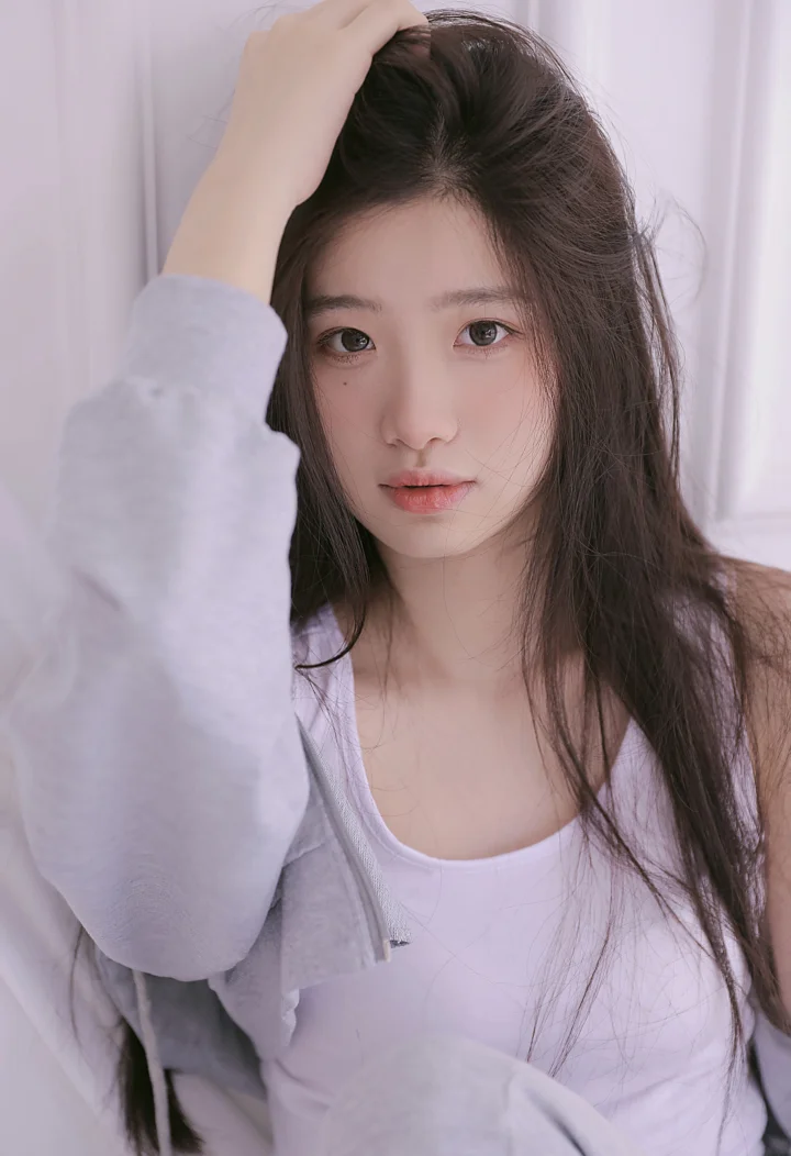 一名长发女性穿着浅色上衣，在室内窗帘前面对镜头摆出肖像姿势。