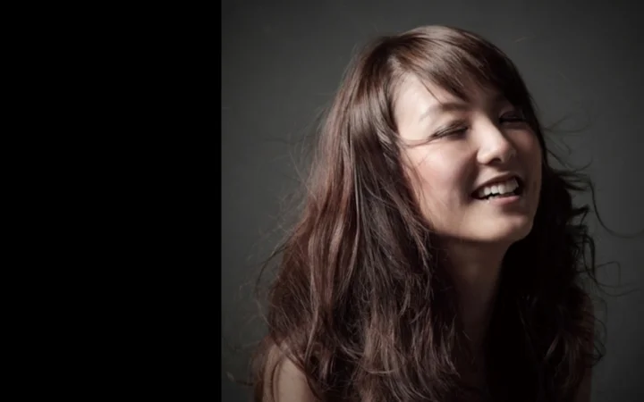 어두운 배경 앞에서 긴 머리의 여성이 미소 짓고 있다.