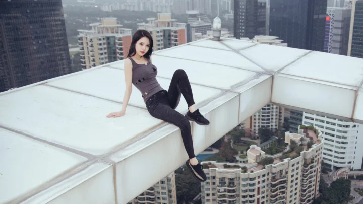 一名成年女性坐在高楼白色屋顶结构上，背景是密集的城市建筑。