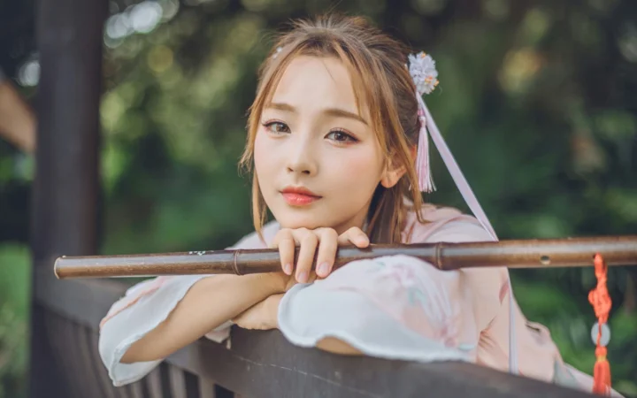 一名身穿浅色传统服饰的女子在户外倚靠木栏，背景是朦胧的绿色树木。