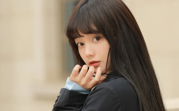 긴 머리의 여성이 손가락을 턱 가까이에 대고 부드럽게 흐려진 배경에서 카메라를 바라보고 있다.