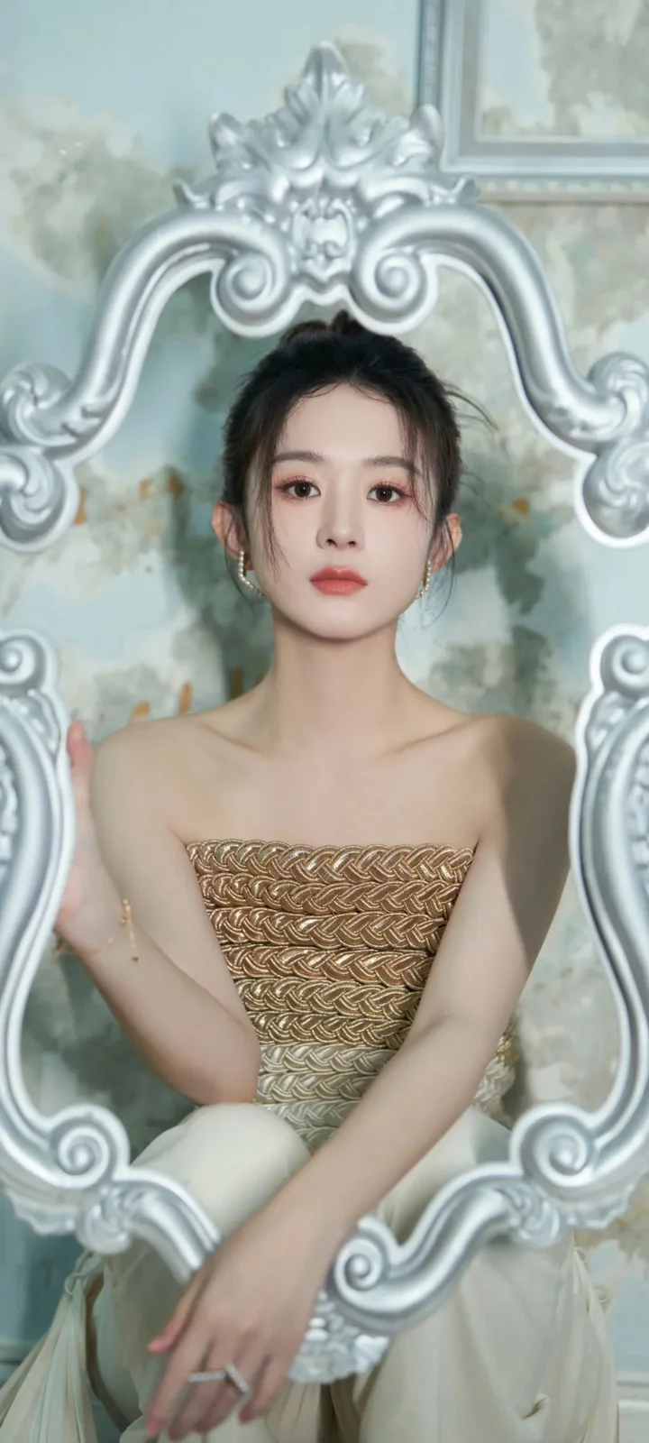 一名成年女性坐在华丽的银色装饰镜框前，穿着金色无肩带礼服。