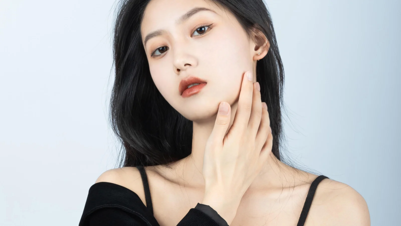 연한 파란색 배경 앞에서 검은색 상의를 입은 검은 머리 여성이 뺨에 손을 대고 있다.