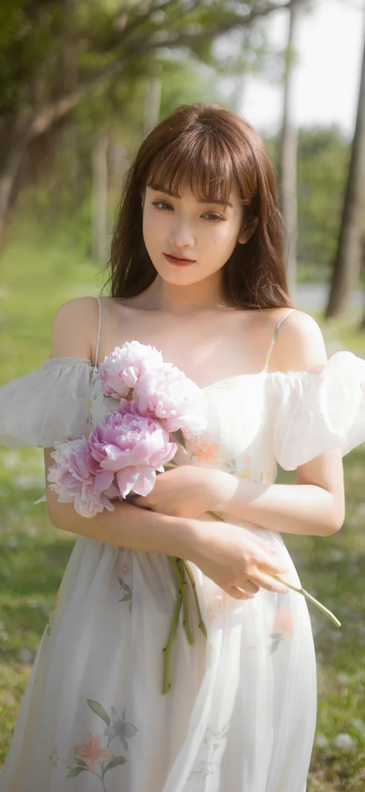 一名成年女子在户外手持粉色花束，身穿白色连衣裙站在阳光下。