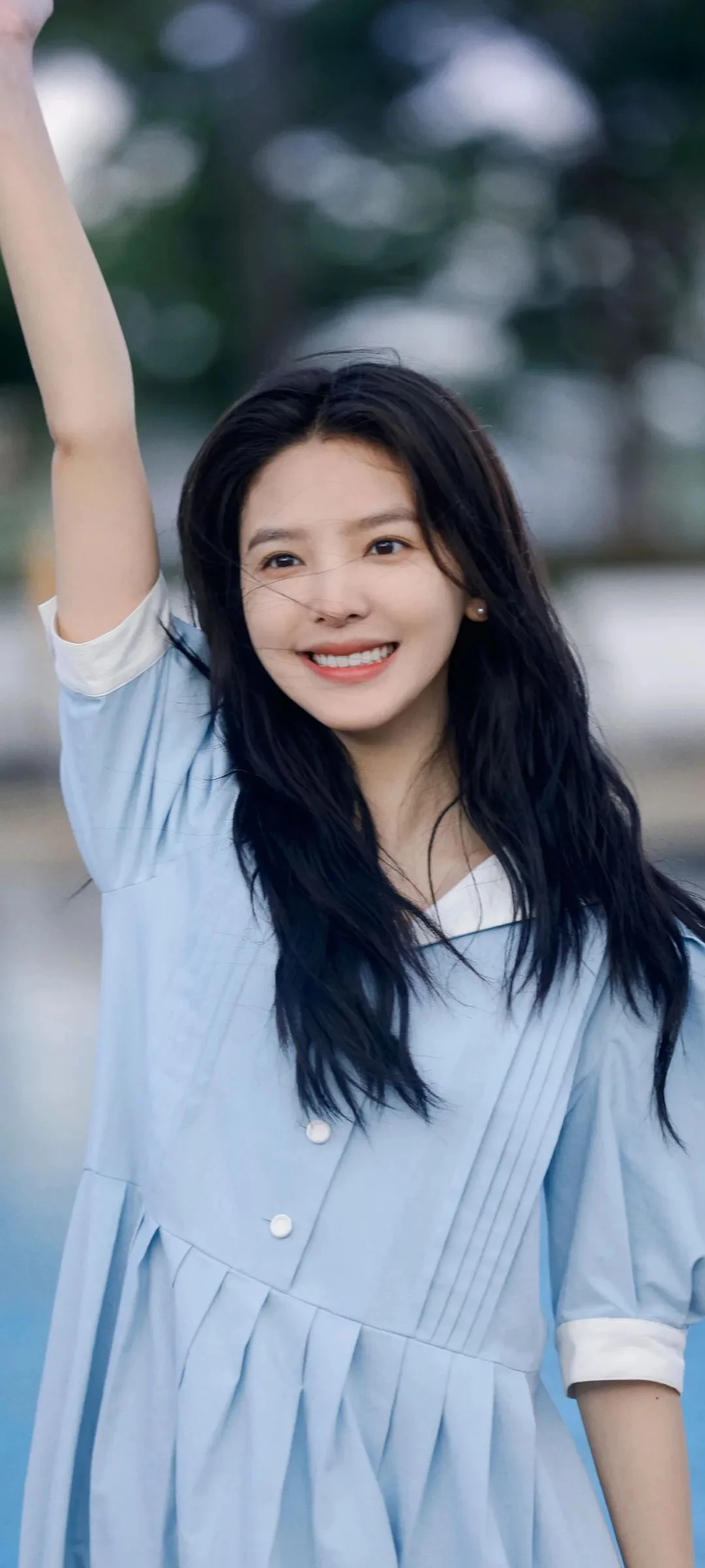 긴 머리의 여성이 야외에서 연한 파란색 옷을 입고 한쪽 팔을 올린 채 미소 짓고 있다.