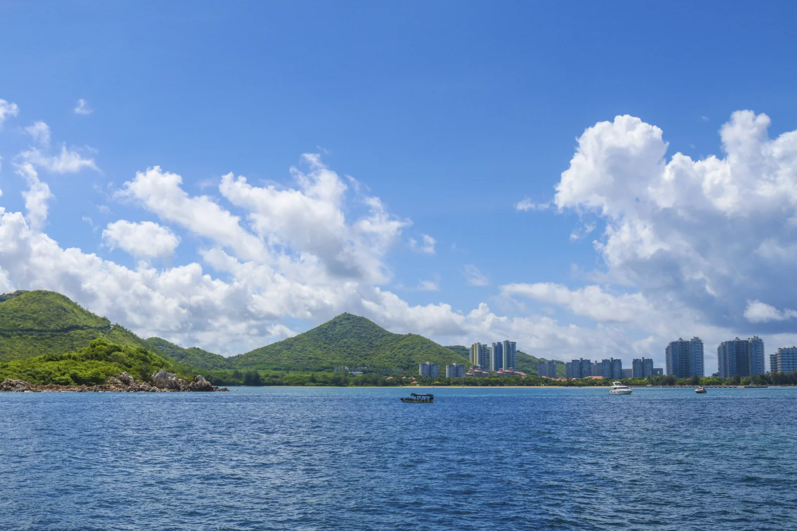 蓝天下的海面延伸至绿色群山与远处城市天际线，白云点缀其间。
