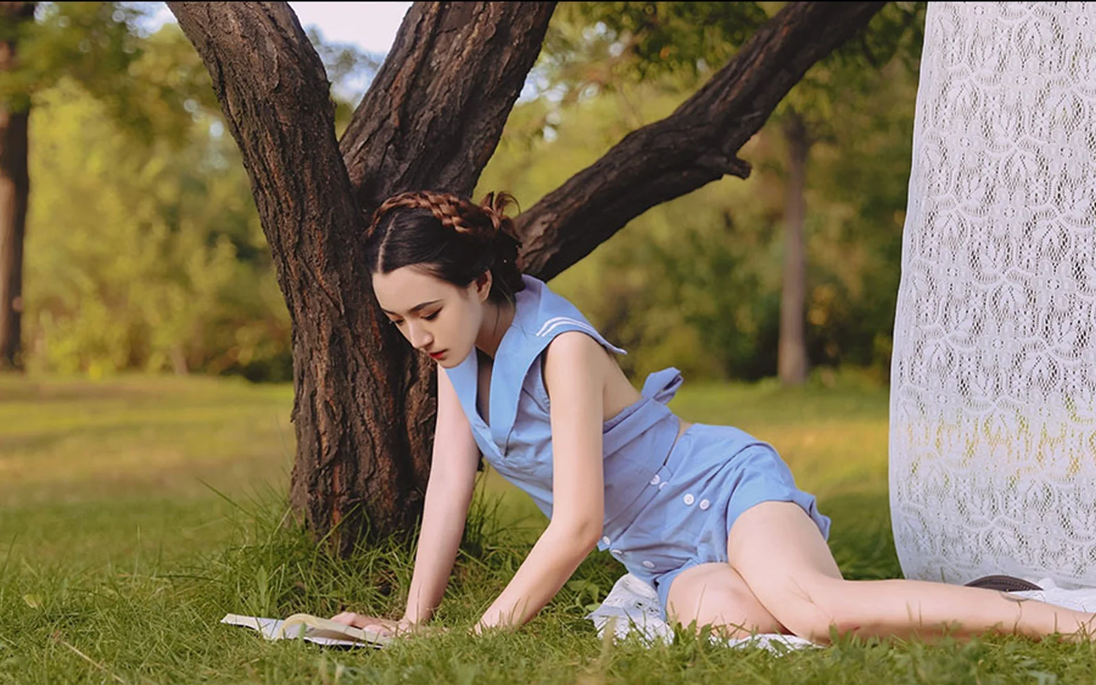 一名身穿浅蓝色连衣裙的成年女子在草地上的树旁俯身。