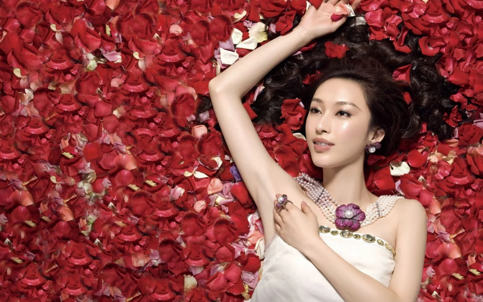 一名成年女性躺在红色玫瑰花瓣上，身穿白色无肩带礼服并佩戴装饰性项链，一只手臂抬起。