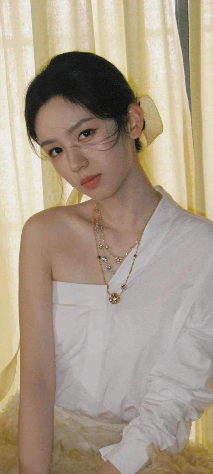 一名女性身穿白色单肩服装，站在浅色窗帘前看向镜头。