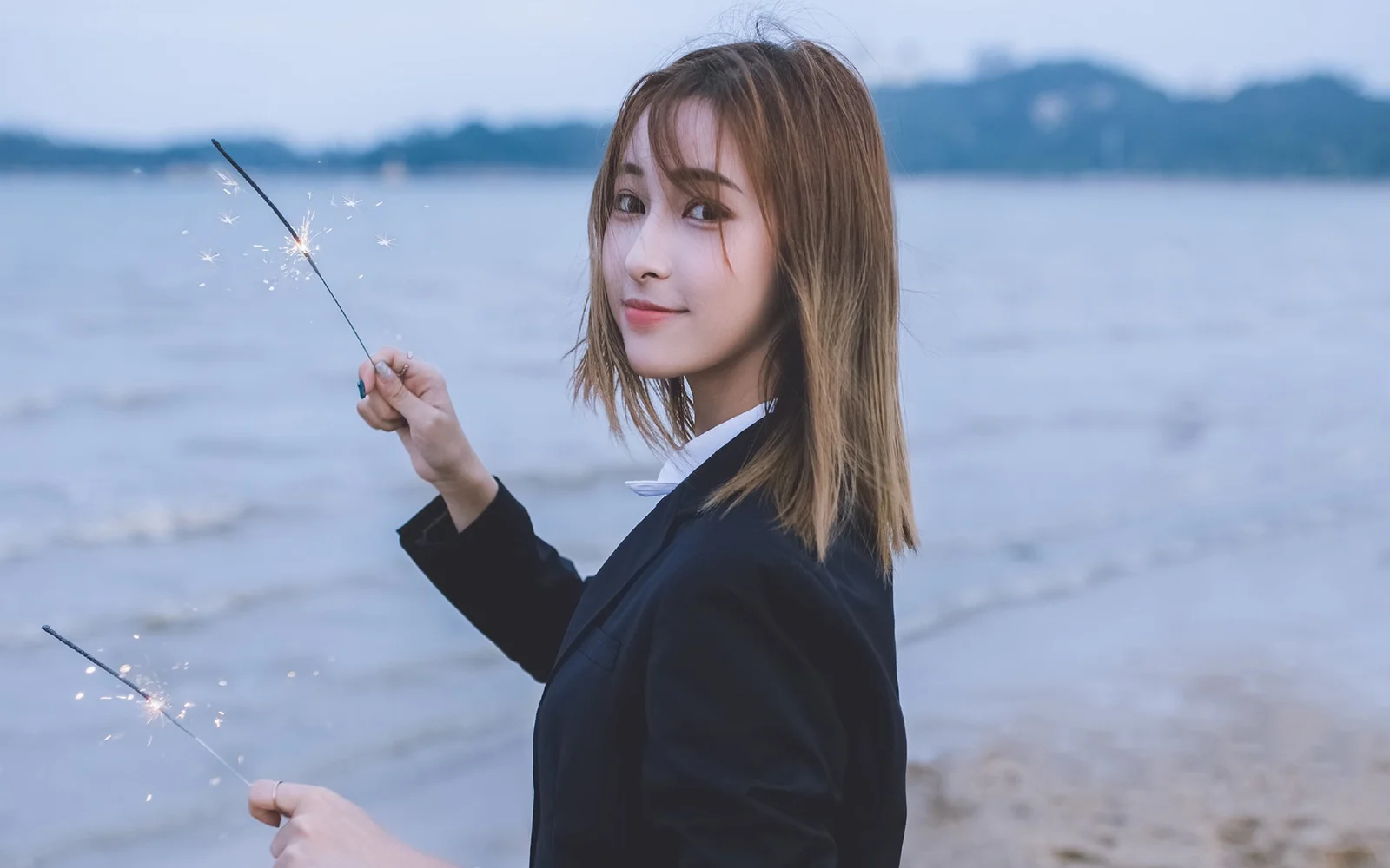 一名女子站在海邊，手持點燃的仙女棒，背景是平靜的海面與遠處的海岸線。