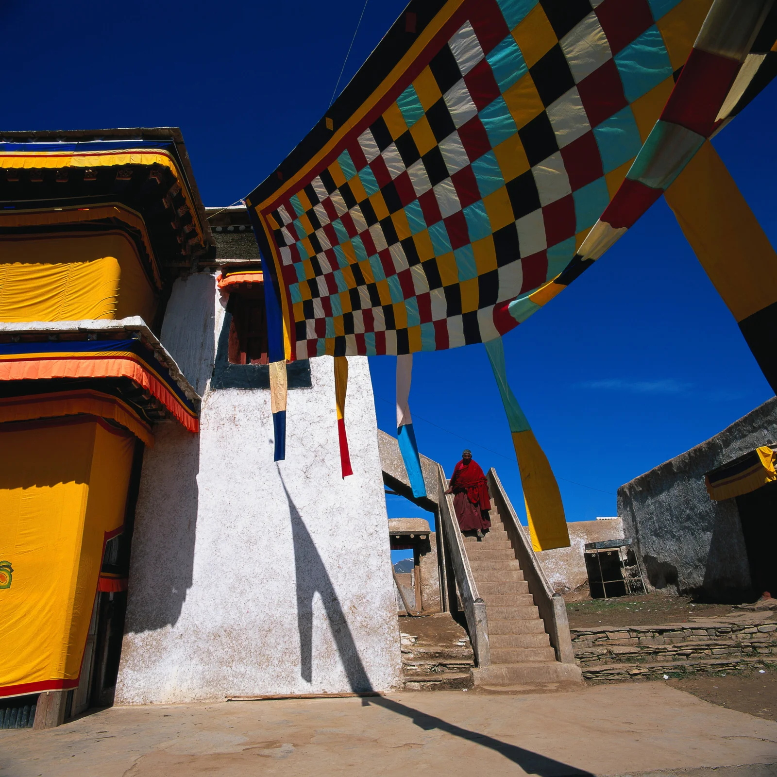 彩色经幡悬挂在寺院建筑上方，蓝天下一名身穿红色衣物的人走下台阶。