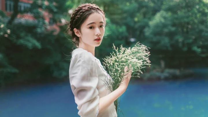 一名女子在树木与蓝色水面前手持一束浅色花枝。