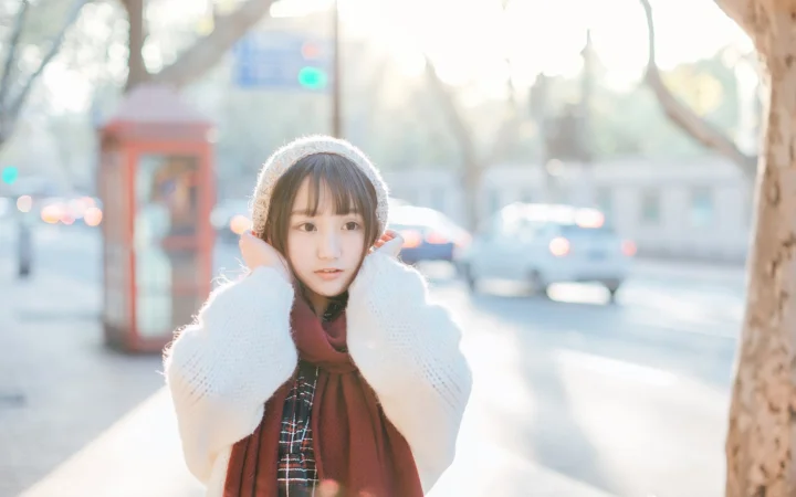 一名戴针织帽、穿白色毛衣并围红色围巾的女性站在阳光明亮的街道旁。