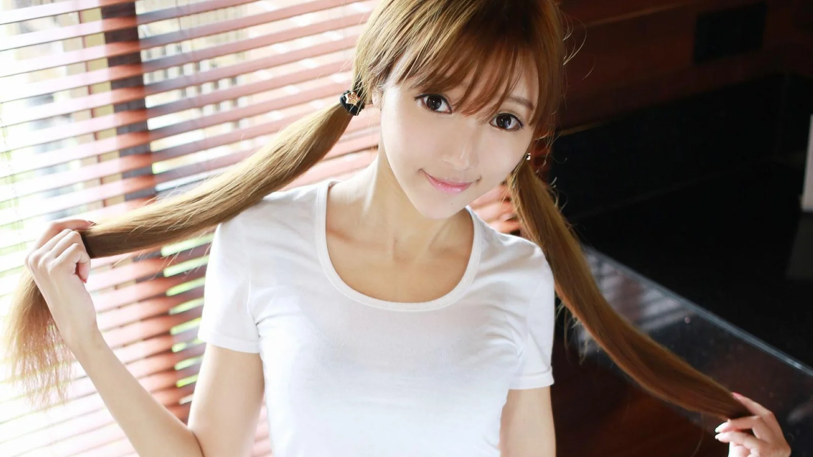 양갈래 머리에 흰색 티셔츠를 입은 성인 여성이 블라인드 옆에서 카메라를 향해 미소 짓고 있습니다.